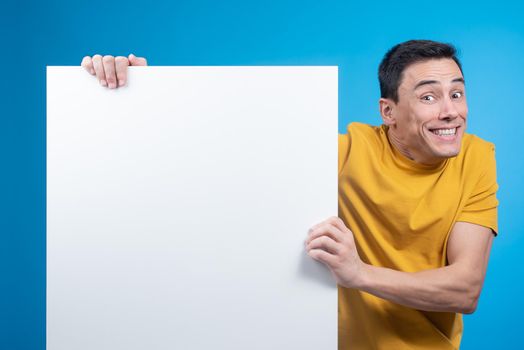 Cheerful young male in yellow t shirt showing empty white poster for advertisement and looking at camera, while widening eyes and raising eyebrows on blue background