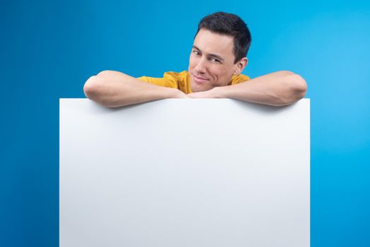 Smiling male model leaning on empty placard for advertisement and looking at camera with tricky face on blue background