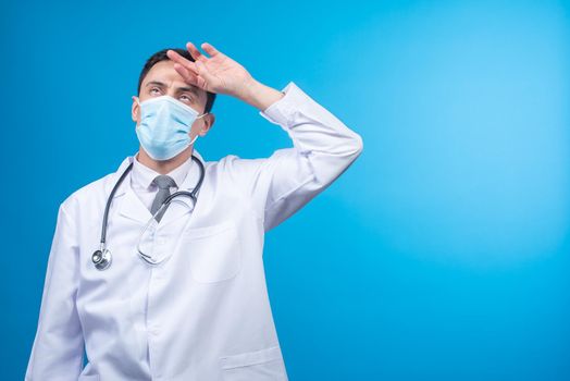 Tired physician in protective mask with stethoscope on neck putting hand on forehead against blue background