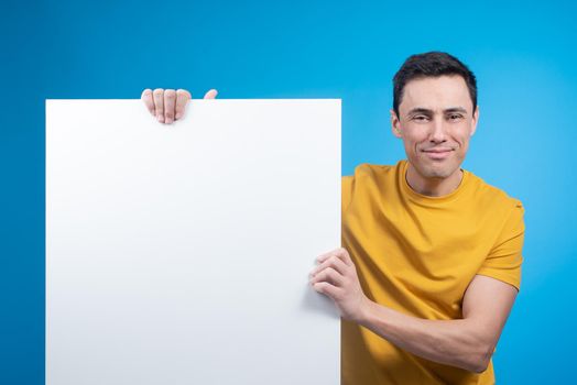 Positive young male in yellow t shirt holding large blank poster and looking at camera on blue backdrop