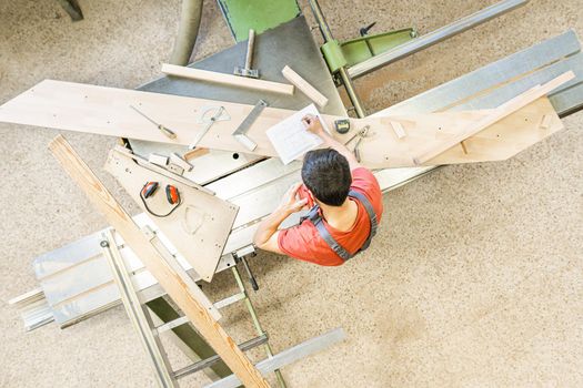 Top view of anonymous male carpenter having phone conversation while taking notes at sliding table saw in professional light joinery