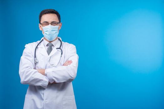 Content physician in protective mask crossing arms and looking at camera with stethoscope on neck on blue backdrop