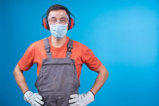 Confident male carpenter in uniform with face mask, goggles, earmuffs and gloves holding hands on waist and looking at camera while standing against blue background during pandemic