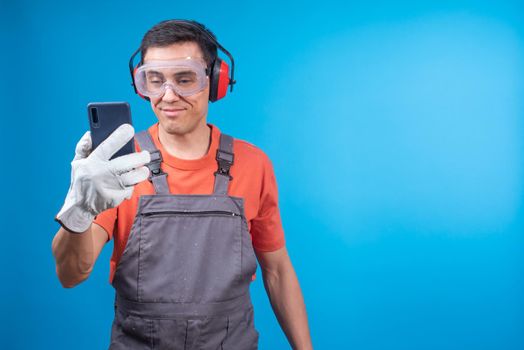 Glad male carpenter in workwear with protective goggles, earmuffs and gloves smiling and checking message in social media on cellphone while taking break in work against blue background