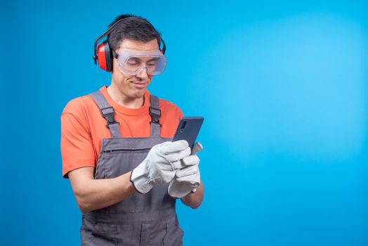 Positive craftsman in uniform with goggles, earmuffs and gloves smiling and texting in social media on mobile phone while taking break in work against blue background