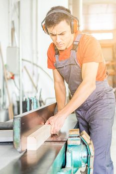 Focused skilled male woodworker in workwear trimming wooden board on special metal woodworking planer while working in light professional carpentry