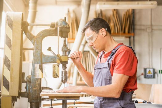 Side view of concentrated male woodworker in protective glasses cutting hole in steel plank while using drilling steel machine in carpentry