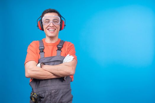 Happy craftsman in workwear with protective goggles and earmuffs crossing arms and laughing at funny joke against blue background