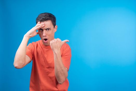 Impressed man in orange t shirt touching forehead and looking at camera with opened mouth while pointing away with thumb against blue background