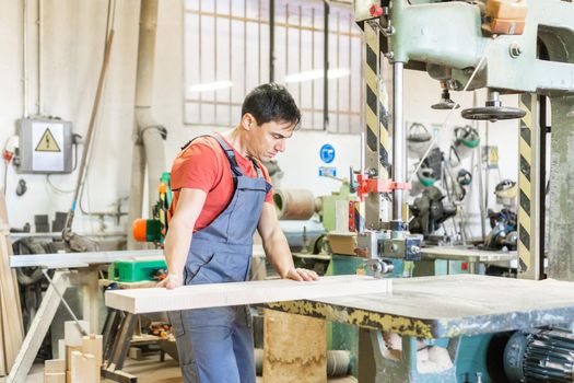 Concentrated male woodworker in workwear cutting long wooden plank with professional metal band saw while working in light carpentry with special equipment