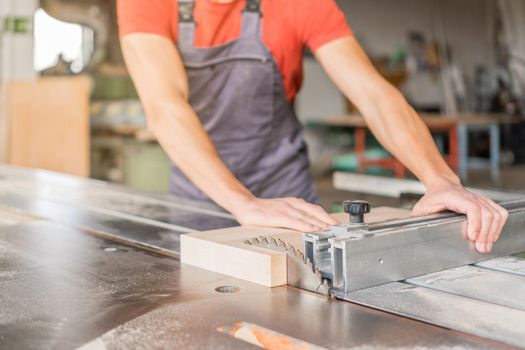 Crop anonymous male master in uniform sawing wooden plank on professional squaring machine while working in light workshop with equipment