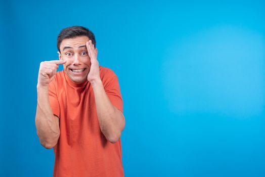 Embarrassed male in orange t shirt hiding face and looking at camera with apologetic smile while pointing away against blue background