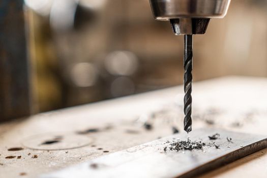 Closeup of metal drill machine drilling hole in steel detail on wooden plank in light professional joinery on blurred background