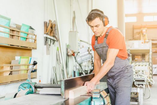 Focused male woodworker in protective headphones and uniform trimming wooden plank on special woodworking planer while working in light joinery