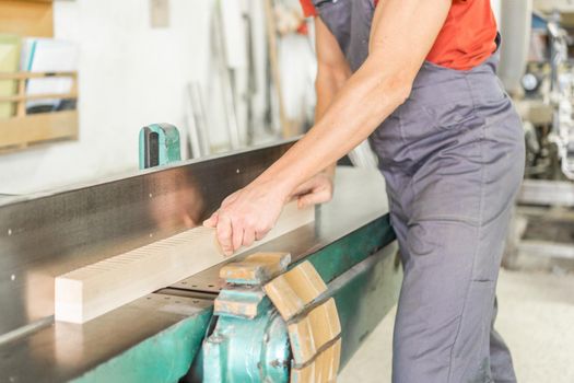 Crop anonymous professional male woodworker in uniform trimming wooden plank on special woodworking planer while working in light spacious carpentry