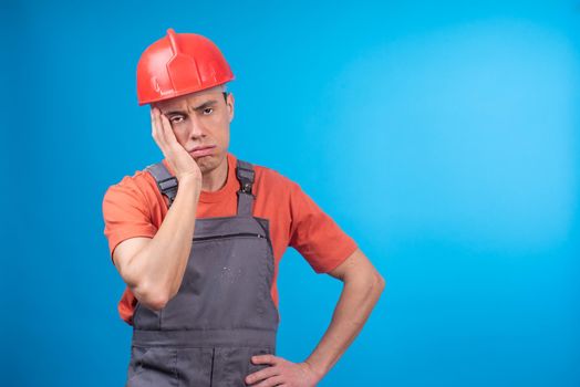 Tired male builder in workwear and hardhat holding hand on waist and touching face while looking at camera in exhaustion against blue background