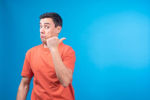 Approving male in orange t shirt looking at camera and pointing away during advertisement campaign against blue background