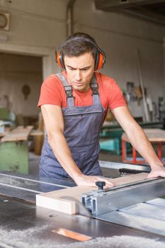 Concentrated male carpenter in protective headphones and uniform cutting wooden plank at squaring machine while working in light professional joinery