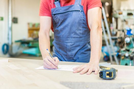 Crop anonymous male woodworker in uniform writing on paper while standing at workbench with measuring tape during work in professional carpentry