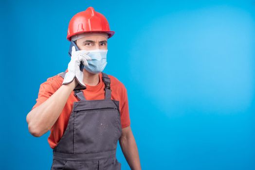 Male contractor in workwear and gloves with face mask and hardhat answering phone call while standing against blue background during pandemic