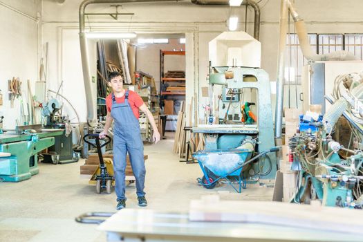 Full body of male worker in uniform pulling pallet truck with wooden boards during work in light professional joinery with various equipment