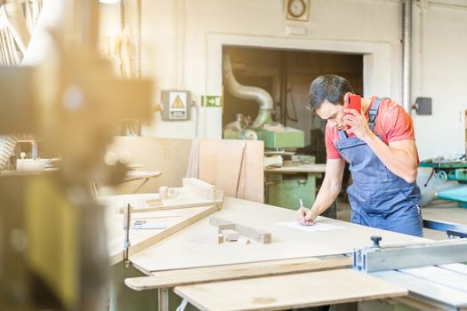 Side view of focused male woodworker having phone conversation while writing at workbench with various supply during work in carpentry