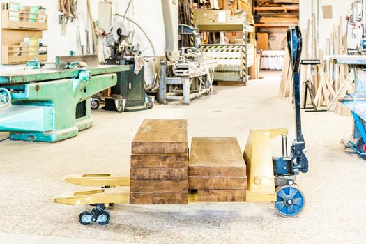 Metal pallet truck with stack of wooden planks placed in light spacious professional carpentry with various special equipment for woodwork