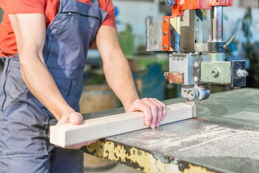 Crop anonymous male master in blue uniform cutting wooden plank with band saw while working in professional joinery