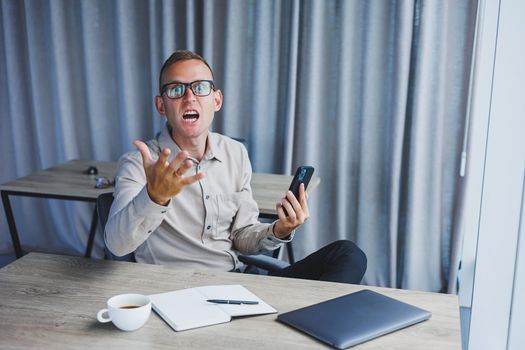 A young attractive male freelancer in glasses sits in the office and looks at the phone. Handsome blogger writing ideas on phone while sitting at wooden table in office with laptop