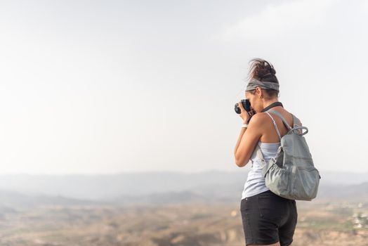 Back view of unrecognizable female traveler with backpack taking picture of mountainous landscape using photo camera while exploring highlands during vacation in summer