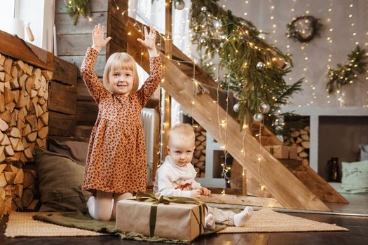 Little brother and sister play on Christmas eve in a beautiful house decorated for the New Year holidays. Children are playing with a Christmas gift. Scandinavian-style interior with live fir trees and a wooden staircase.