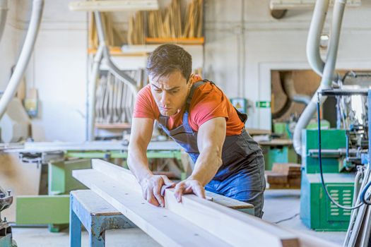 Concentrated male craftsman in uniform smoothing piece of wood while working in light workshop