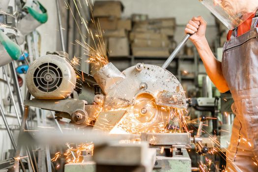 Crop unrecognizable artisan in protective mask and uniform cutting steel with sparks on special equipment in carpentry studio