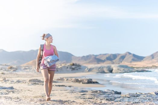 Full body of slim barefoot female traveler in summer clothes and with backpack walking along sandy seashore, while enjoying vacation in sunny summer day in Almeria Andalusia Spain