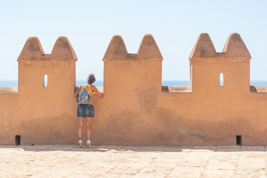 Back view of unrecognizable woman with backpack admiring sea view, while standing near ancient stone towers of Citadel of Almeria city in Andalusia Spain in sunny day