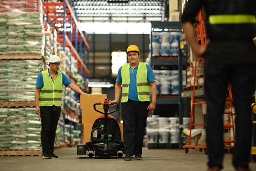 Shot of warehouse workers are pulling a pallet truck walking through rows of tall shelves full of packed boxes. Logistic industry concept.