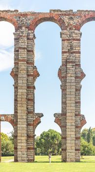 Anonymous traveler standing on grassy ground under aged columns of Roman aqueduct ruins against green trees in sunlight in Spain