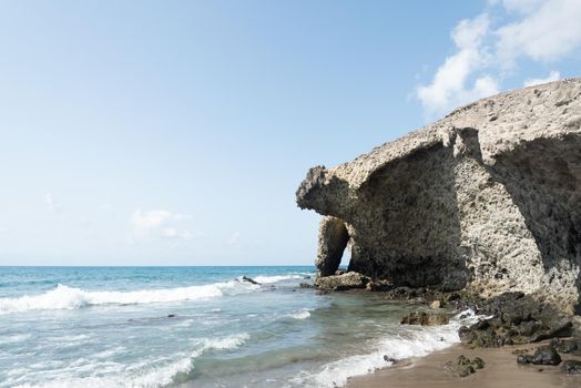 Scenic seascape of blue endless wavy foamy sea washing rough rocky cliff near sandy coast under blue cloudy sky in Almeria in Andalusia in Spain