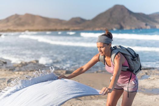 Side view of slim woman in shorts and top with backpack putting towel on sandy beach near waving sea water, and rocky hills during summer vacation in Almeria Andalusia Spain