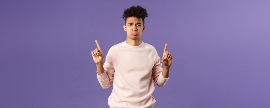 Portrait of gloomy, pouting frowning hipster guy dont have something he wants, pointing fingers up at super cool expensive thing, asking for it, trying receive compassion, purple background.