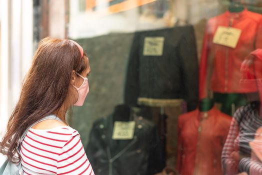 Side view of unrecognizable woman in protective mask looking at clothes on showcase behind glass in shop while walking on street in city