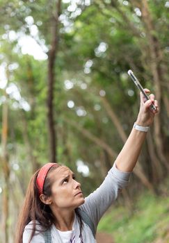 Frowning adult Hispanic female traveler trying to catch mobile connation while browsing cellphone in green forest in Spain and looking up