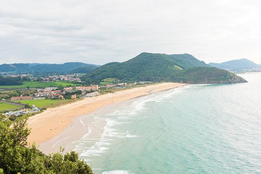 Amazing scenery of sandy seashore with hills covered with green trees located near settlement under cloudy sky in summer day