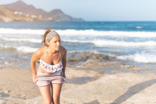 Slim female traveler wearing bikini taking off shorts and looking away, while resting on sandy seashore near waving water and hills during summer vacation in Almeria Andalusia Spain