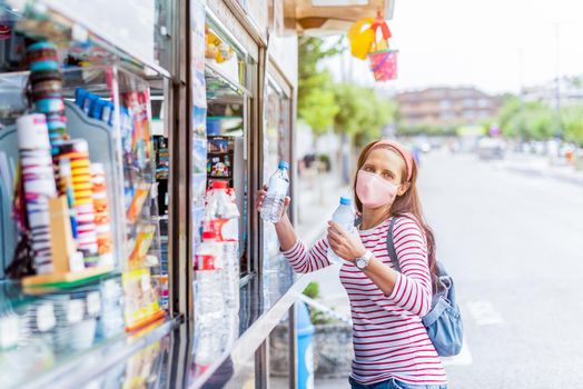 Hispanic female tourist with backpack wearing protective mask buying bottles of water in street stall while enjoying weekend in city