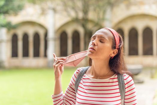 Carefree ethnic female tourist with closed eyes enjoying summer holidays in Spain and taking off mask from face while walking in sunny day