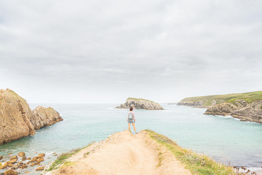 Back view of unrecognizable woman with backpack admiring view of blue sea, while standing on sandy coast near rocky formation on Arnia beach in Cantabria in Spain in