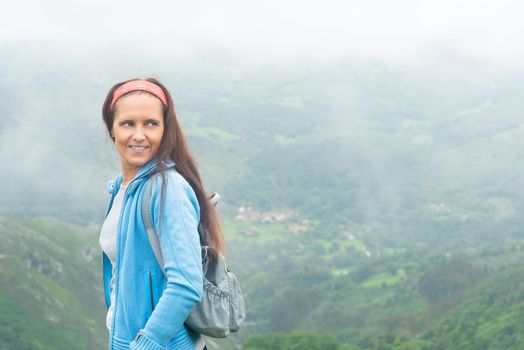 Side view of delighted Hispanic woman with backpack enjoying vacation while standing on edge of mountain against misty landscape of green valley and looking away