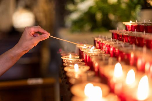 Crop anonymous female lighting flaming candle near many burning candles with wooden stick during praying in church