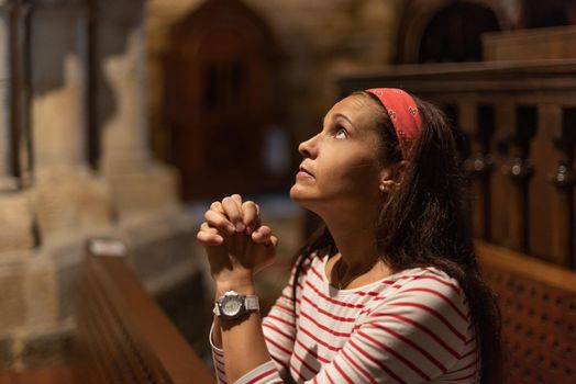 Side view of serious Hispanic female with long dark hair giving prayer with clasped hands, while sitting on bench in old Catholic church and looking up